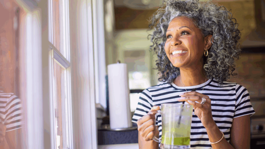 Smiling midlife woman standing by a window holding a glass, enjoying a quiet moment and embracing a peaceful new chapter in life.