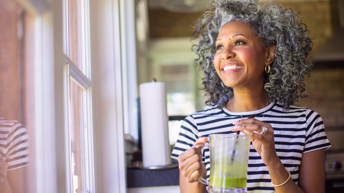 Smiling midlife woman standing by a window holding a glass, enjoying a quiet moment and embracing a peaceful new chapter in life.