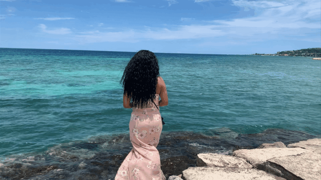 A woman standing at the beach in Jamaica, facing the ocean as the waves roll in, symbolizing peace, reflection, and letting go gracefully.