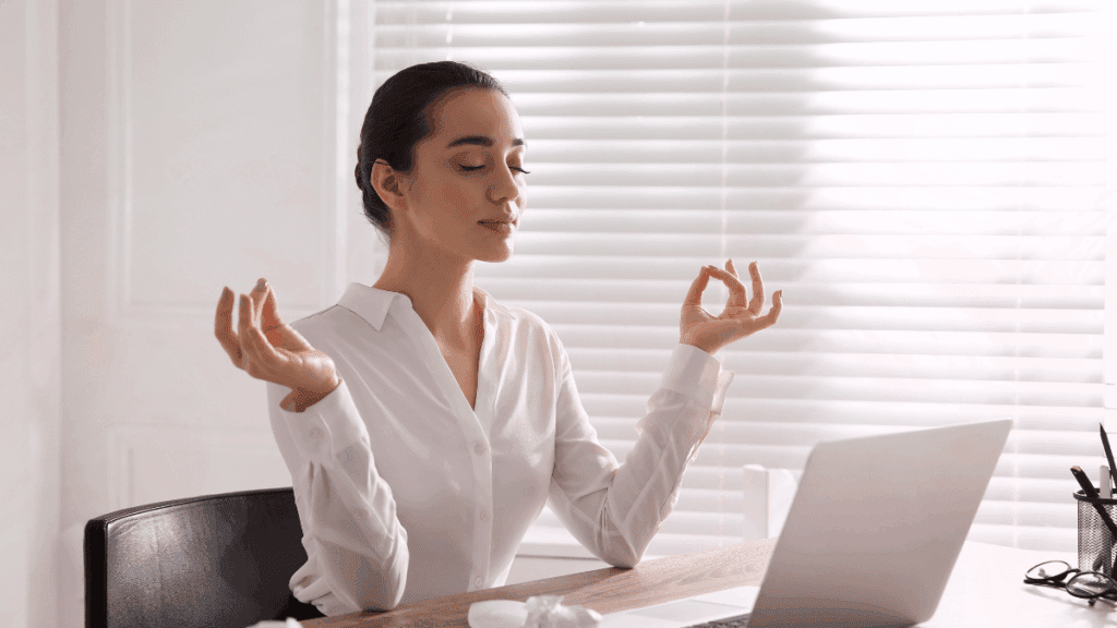 Woman meditating calmly at her desk with soft natural light, symbolizing balance, focus, and inner peace.