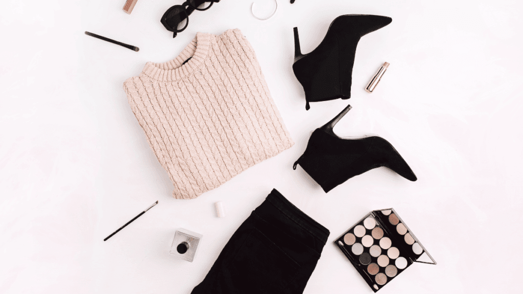 Flat lay of a beige sweater, black jeans, ankle boots, and makeup accessories arranged on a white background, symbolizing modern midlife style and confidence.